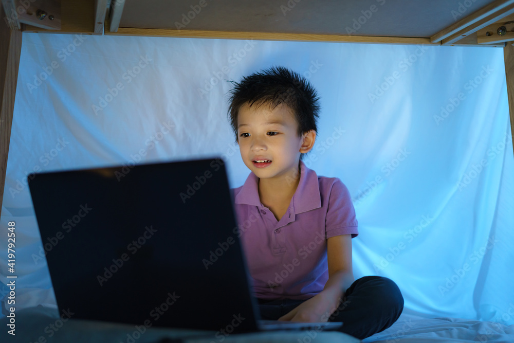 Asian boy child to make a camp to play imaginatively watching a film on laptop in the darkness of th