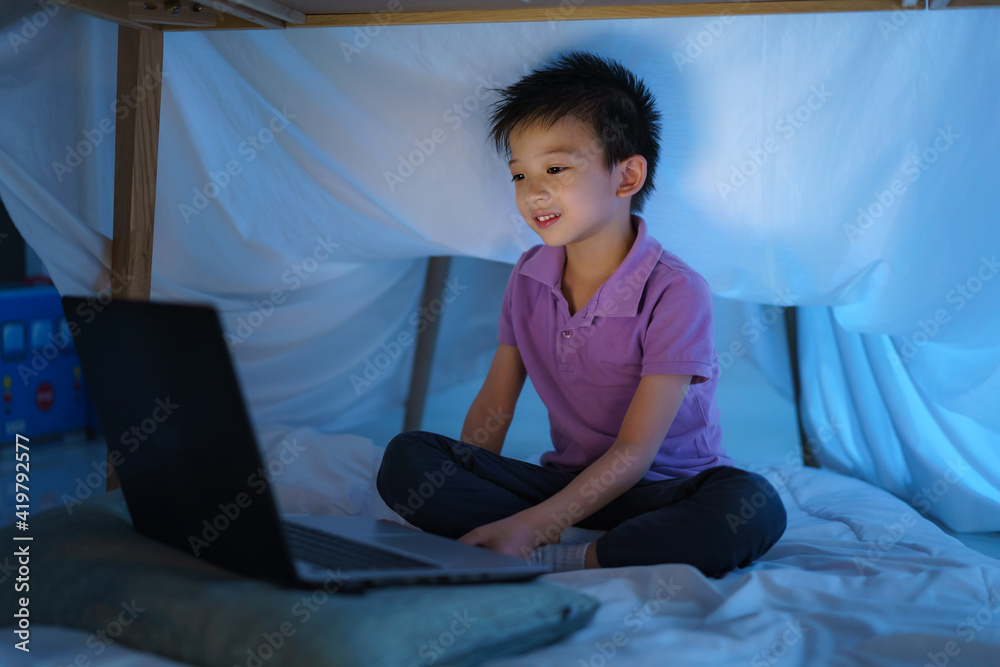 Asian boy child to make a camp to play imaginatively watching a film on laptop in the darkness of th