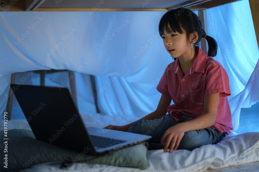 Asian girl child to make a camp to play imaginatively watching a film on laptop in the darkness of t