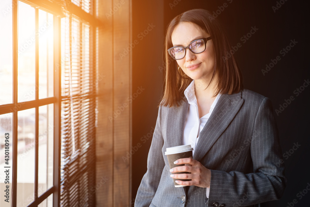 美丽的商务女性，拿着咖啡站在窗户边，阳光明媚。