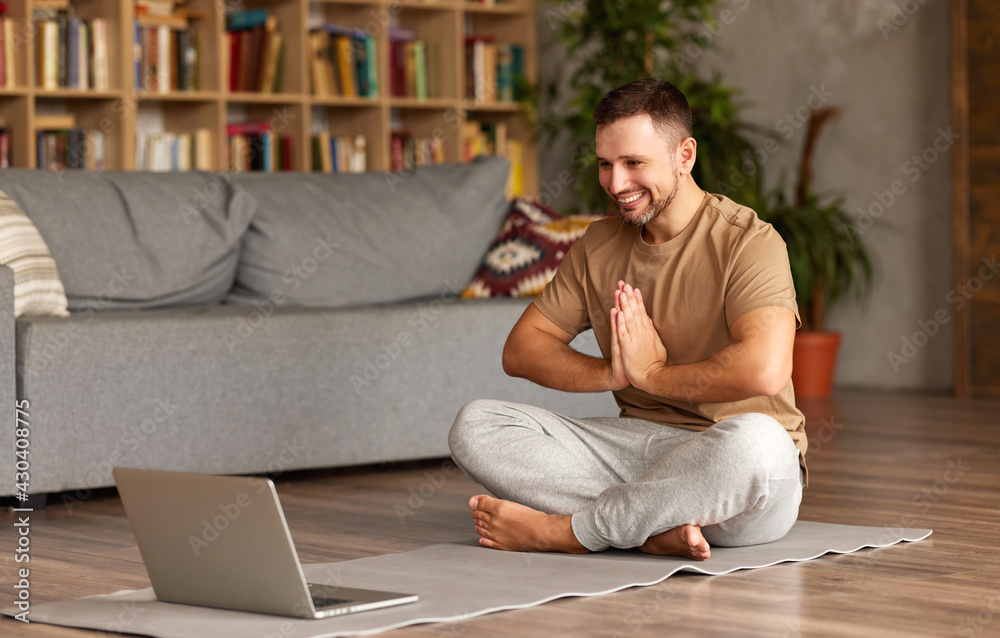 Young cheerful man in casual wear sitting in lotus pose in front of laptop at home