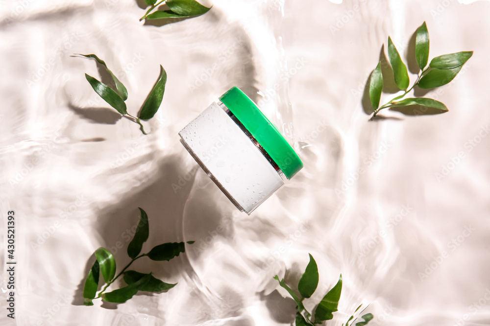 Jar of cosmetic product and leaves in water on light background