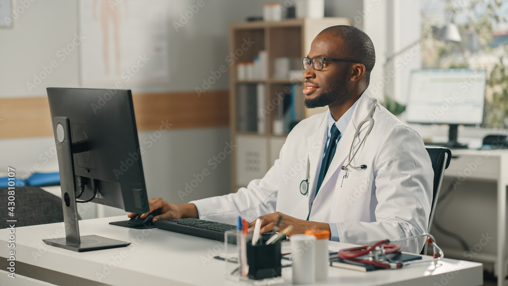 Experienced African American Male Doctor Wearing White Coat Working on Personal Computer at His Offi