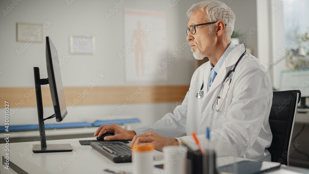 Experienced Middle Aged Male Doctor Wearing White Coat Working on Personal Computer at His Office. S