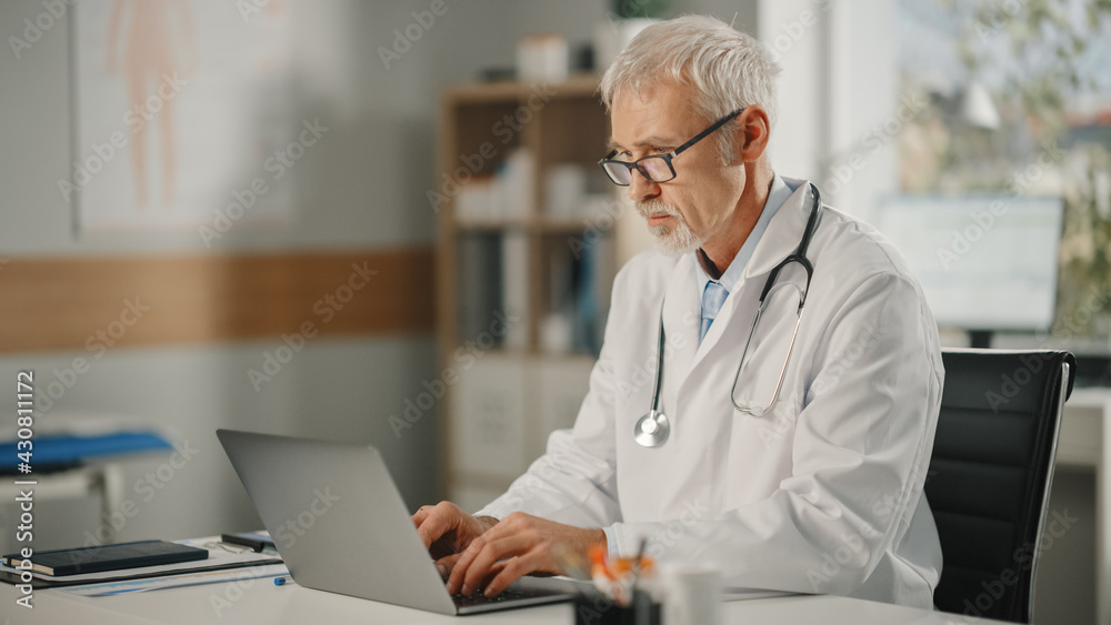 Portrait of an Experienced Middle Aged Male Doctor Wearing White Coat Working on Laptop Computer at 