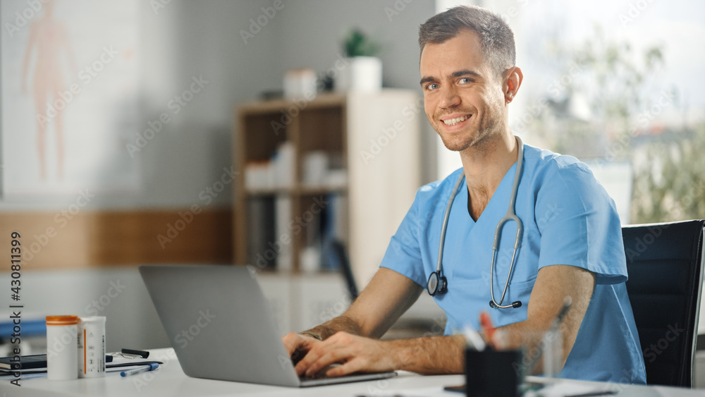 Male Nurse Wearing Blue Uniform Working on Laptop Computer at Doctors Office and Smiling on Camera.