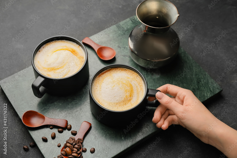 Female hand with cups of hot cappuccino and coffee pot on dark background