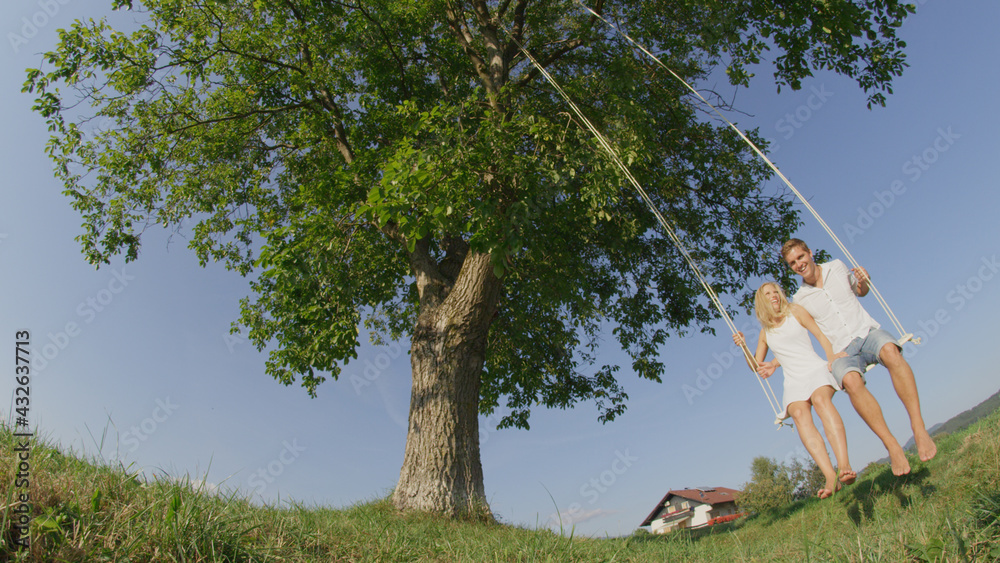 LOW ANGLE: Lovely young couple swaying on swing in green meadow in sunny spring.