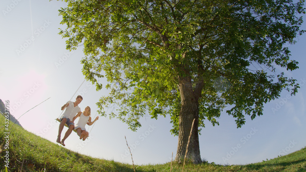 LOW ANGLE LENS FLARE Young couple swinging under big green tree in sunny meadow