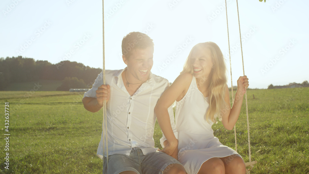 LENS FLARE PORTRAIT Couple laughs heartily while swaying on rope swing in spring