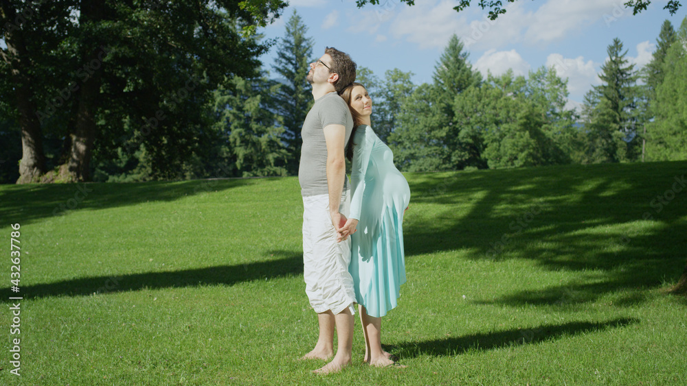 Young pregnant couple standing with their back to each other in a green meadow