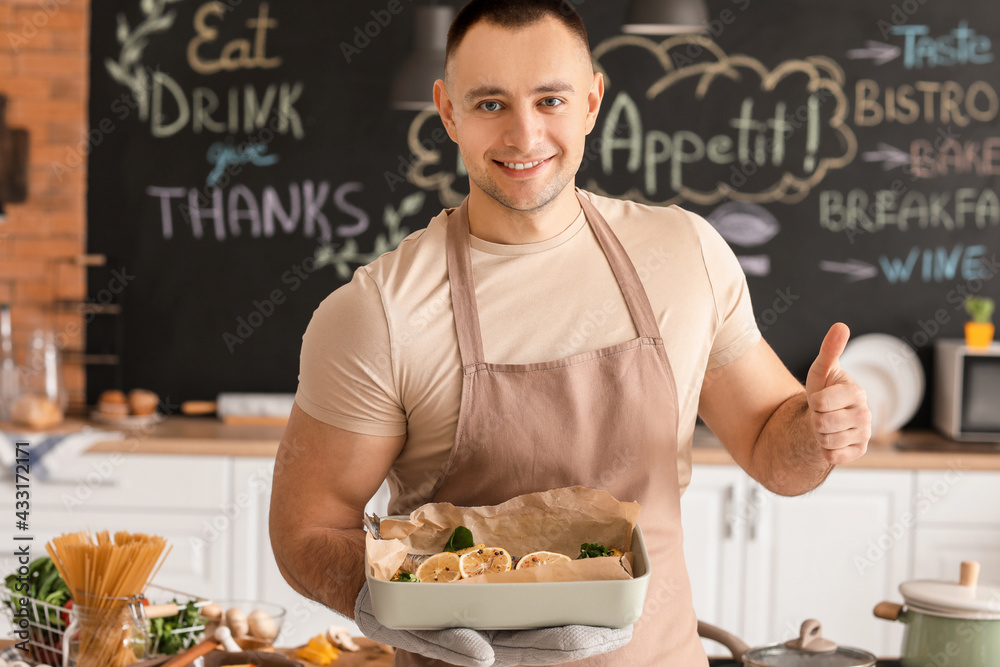 Young man with baking dish showing thumb-up in kitchen