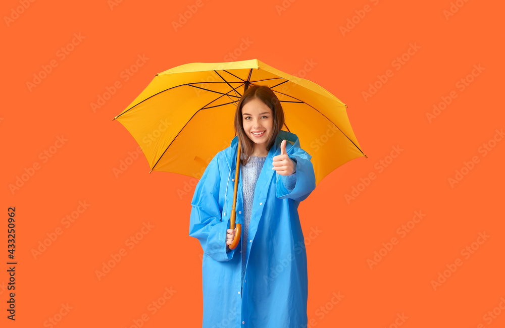 Stylish young woman in raincoat and with umbrella showing thumb-up on color background