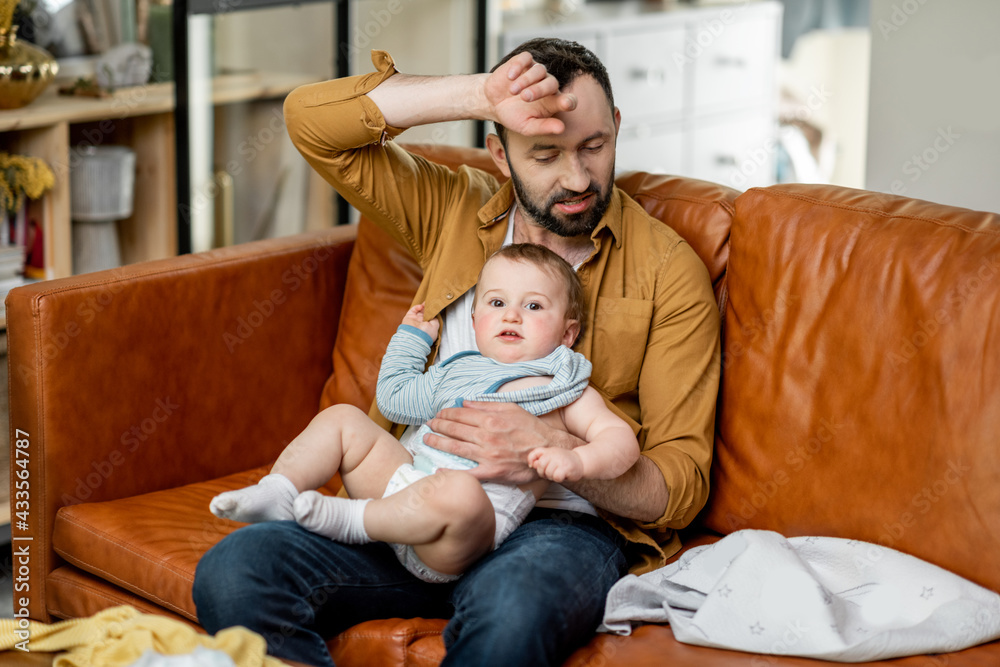 Tired father nursing a newborn child while sitting on sofa at home with scattered kids clothes. Fath