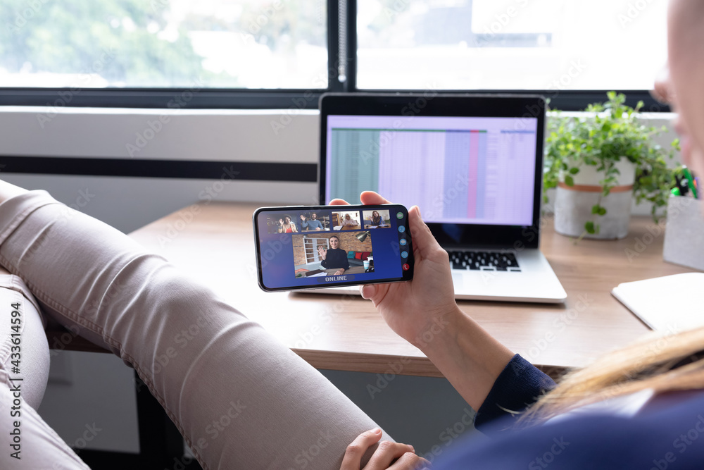 Caucasian businesswoman sitting at desk using smartphone having video call with group of colleagues