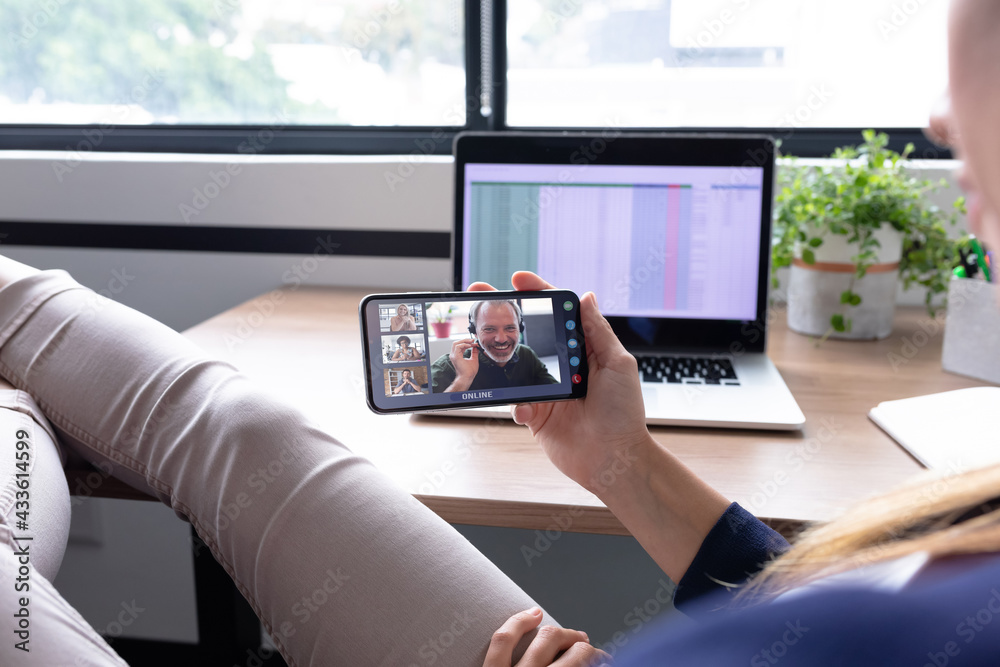 Caucasian businesswoman sitting at desk using smartphone having video call with group of colleagues