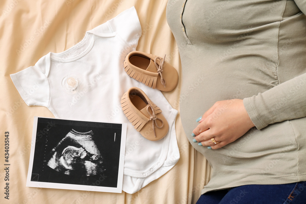 Pregnant woman with sonogram image and baby clothes on bed