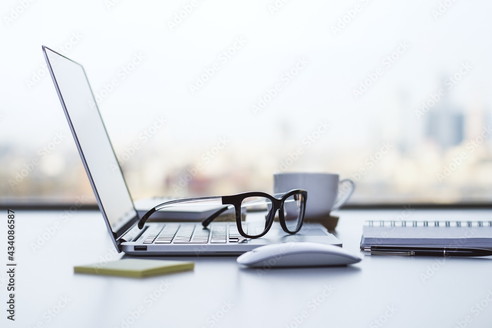 Stylish workspace with white table and laptop, glasses and notebook on it at blurry city background.