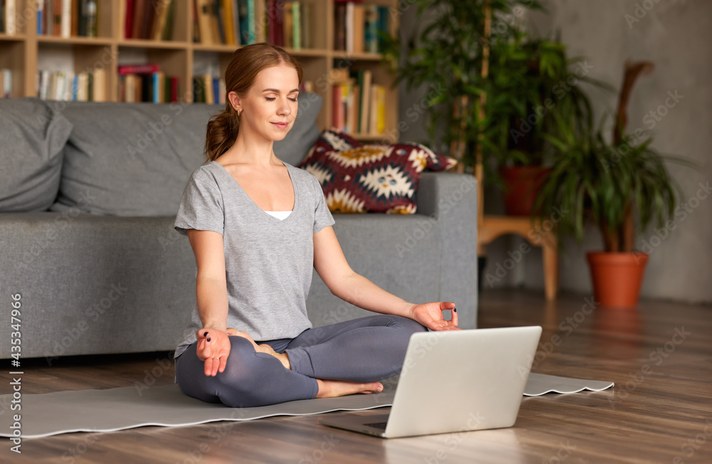 Peaceful young woman meditating while sitting in lotus pose on floor at home in front of laptop