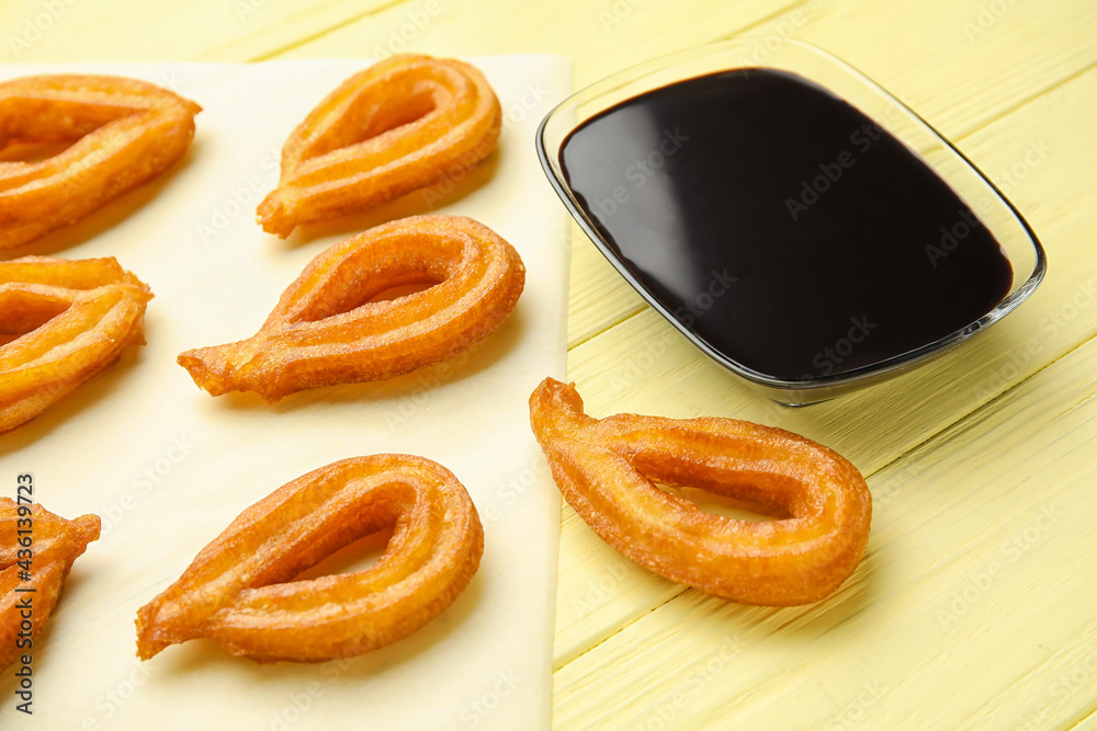 Parchment with tasty churros and melted chocolate sauce on color wooden background, closeup