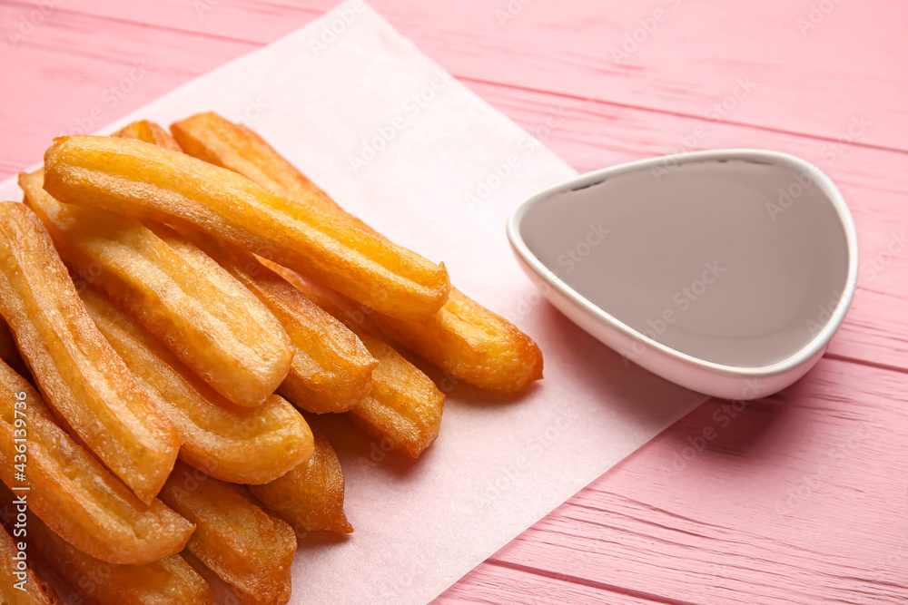 Parchment with tasty churros and melted chocolate sauce on color wooden background, closeup