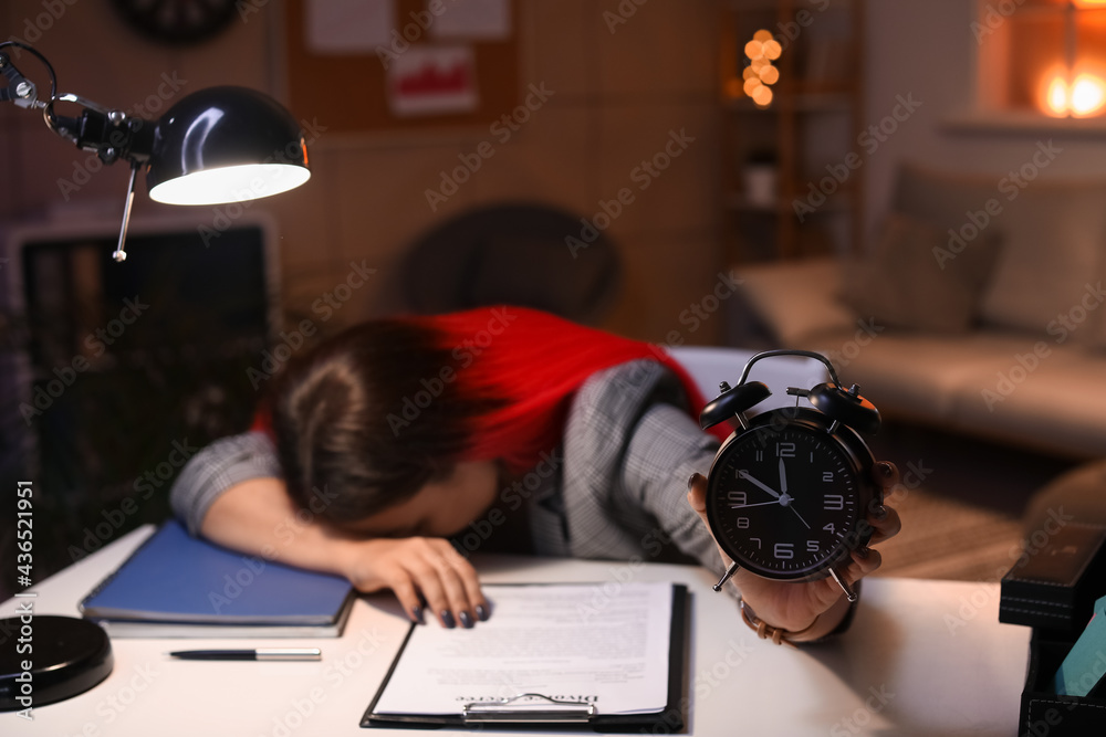 Tired young woman with alarm clock in office at night