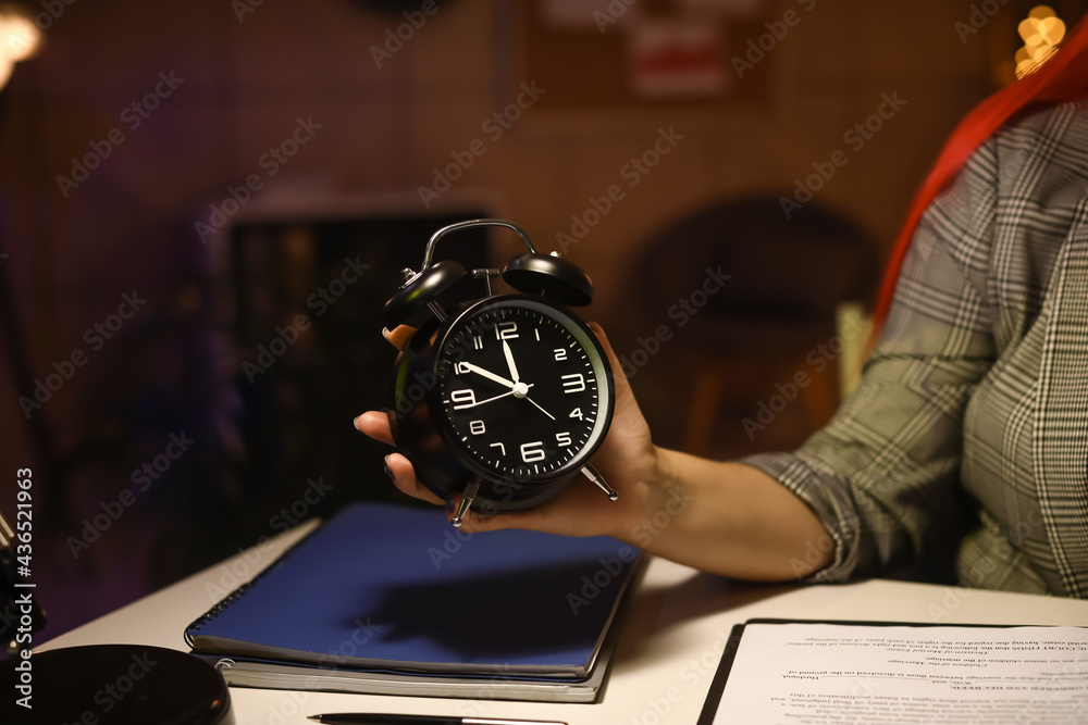 Young woman with alarm clock in office at night