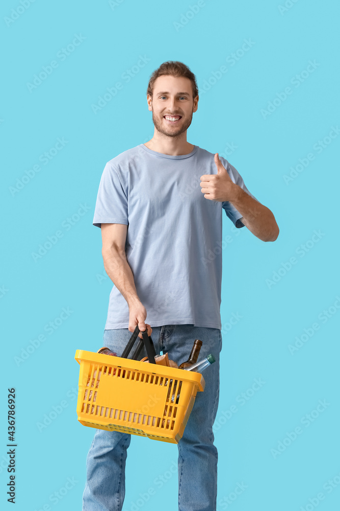 Young man with shopping basket showing thumb-up on color background