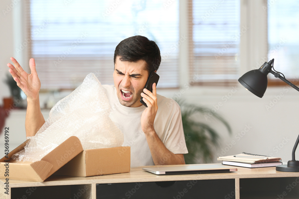 Angry young man with parcel talking by mobile phone at home