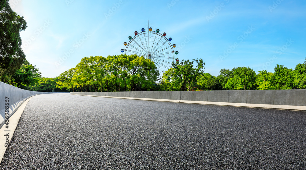 Empty asphalt road and ferris wheel with green forest under blue sky.