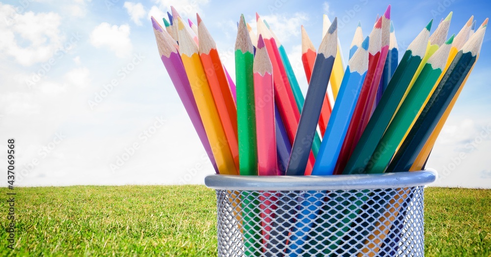 Composition of coloured pencils in mesh metal container, in sunny field with blue sky