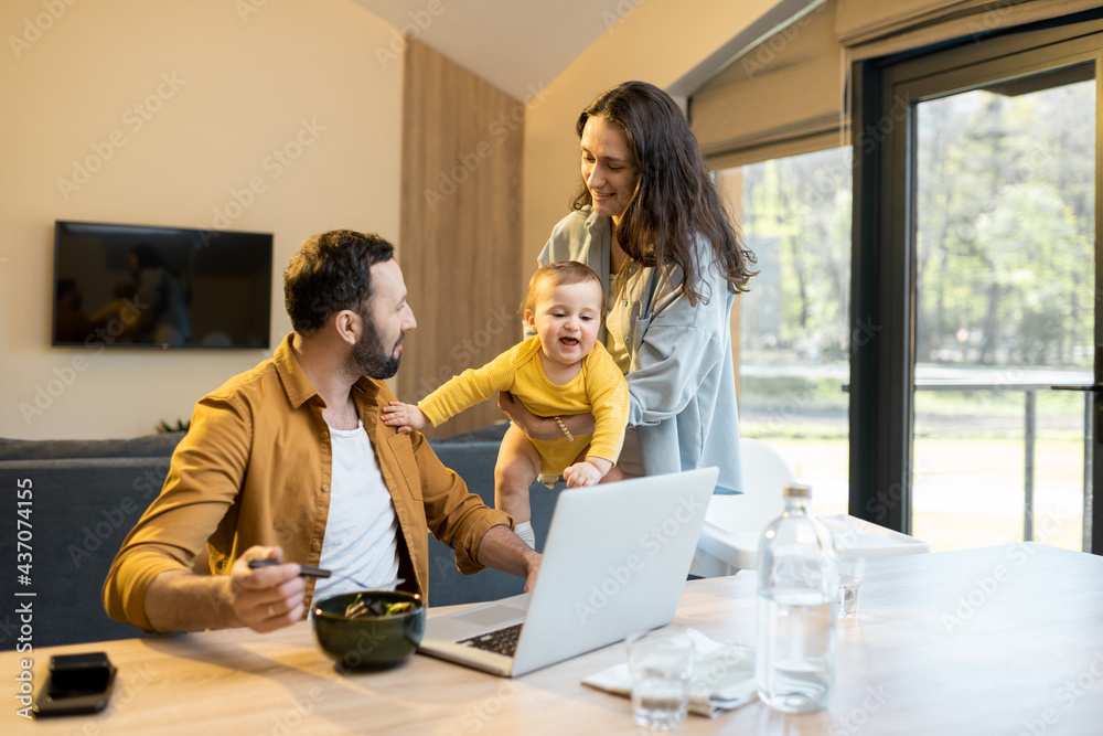 Young father works on a laptop during a lunch time at home with his wife and one year son. Home fami