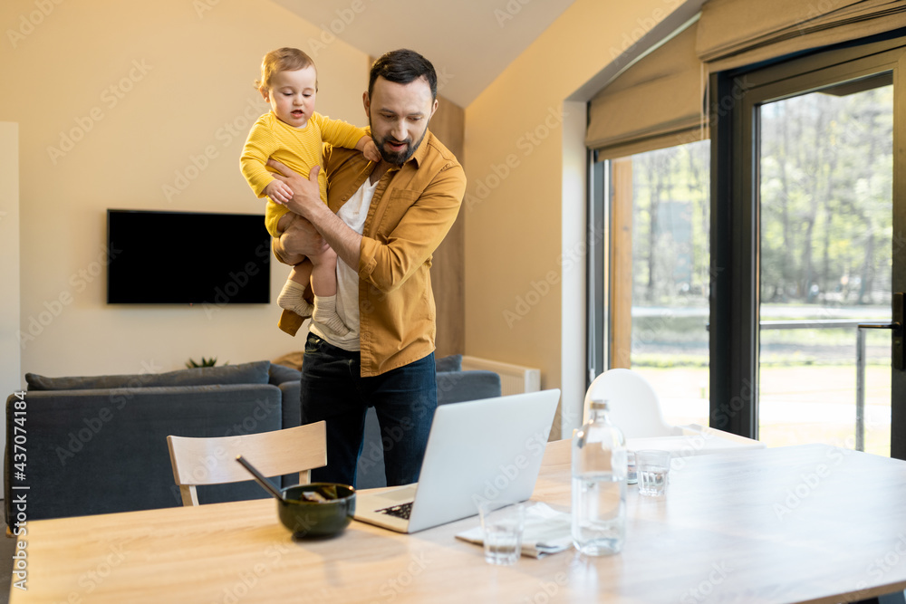 Multitasked father nursing his baby son and works on a laptop during a lunch time at home. Fatherhoo