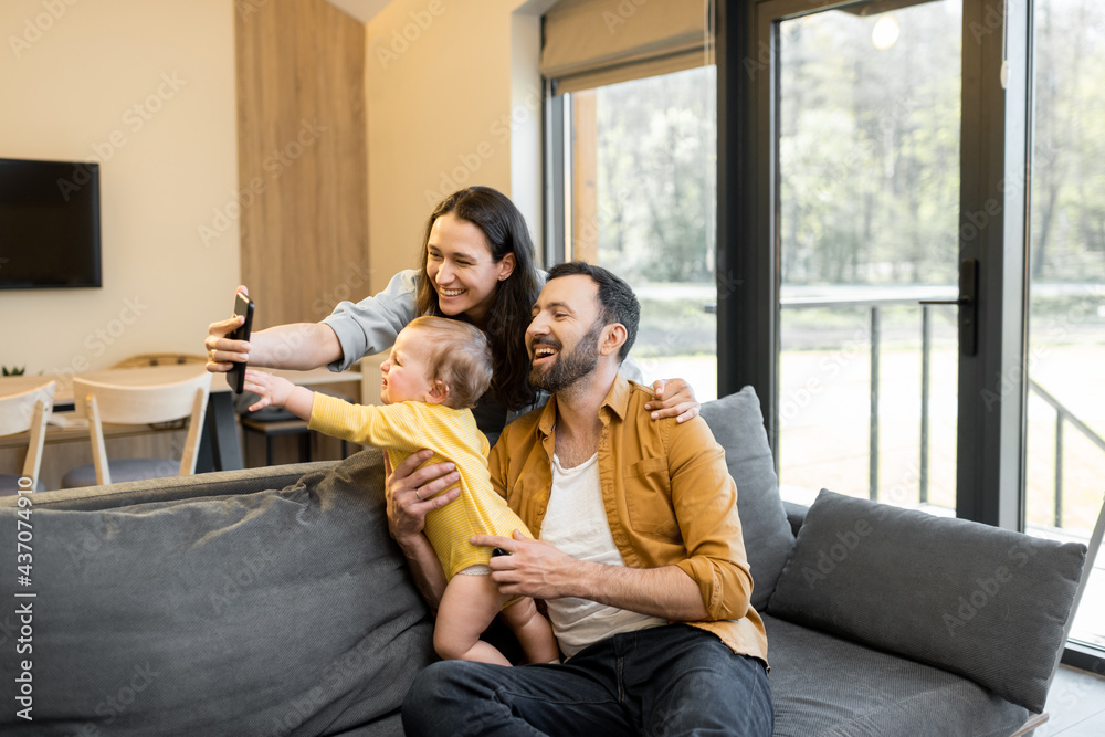 Happy family with a one year baby boy having a video call on phone from home. Concept of remote fami