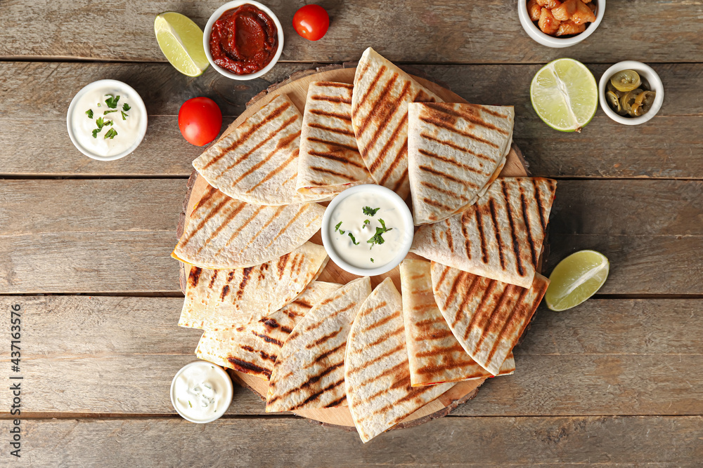 Board with tasty quesadillas, lime and sauces on wooden background