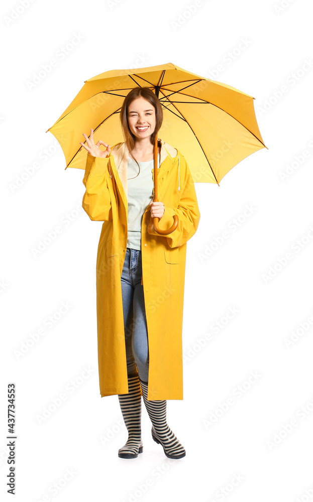 Stylish young woman in raincoat and with umbrella showing OK on white background