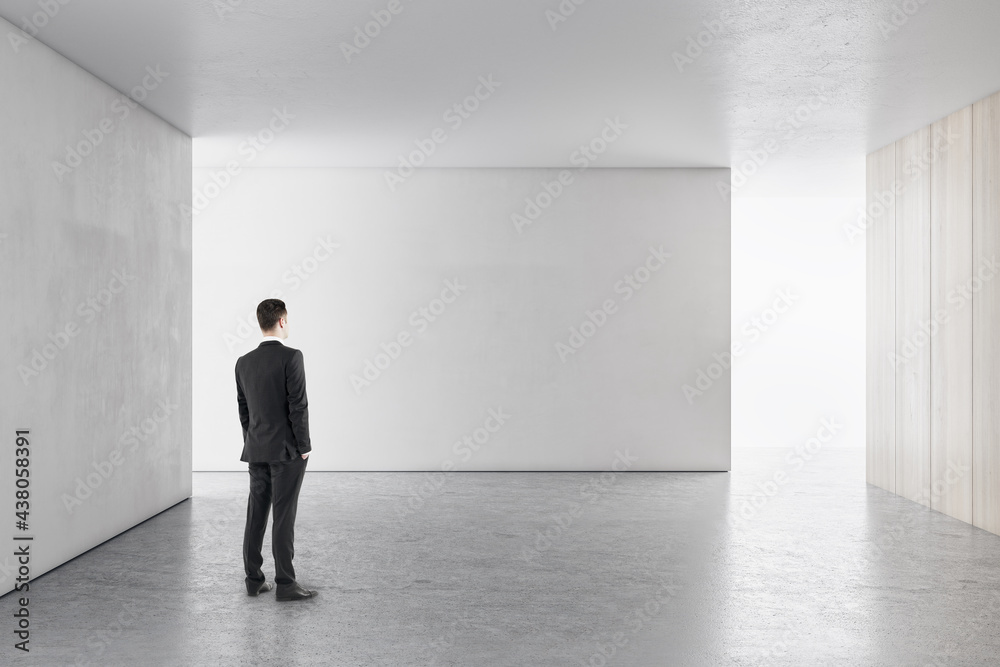 Businessman in bright concrete interior with empty wall looking at mockup place for your advertiseme