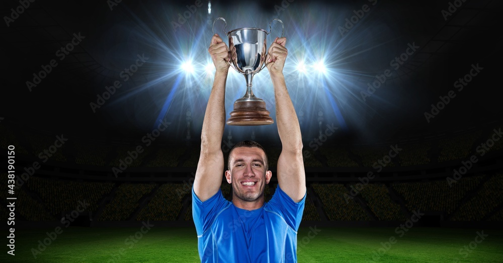 Portrait of caucasian male athlete lifting a trophy against floodlights in background