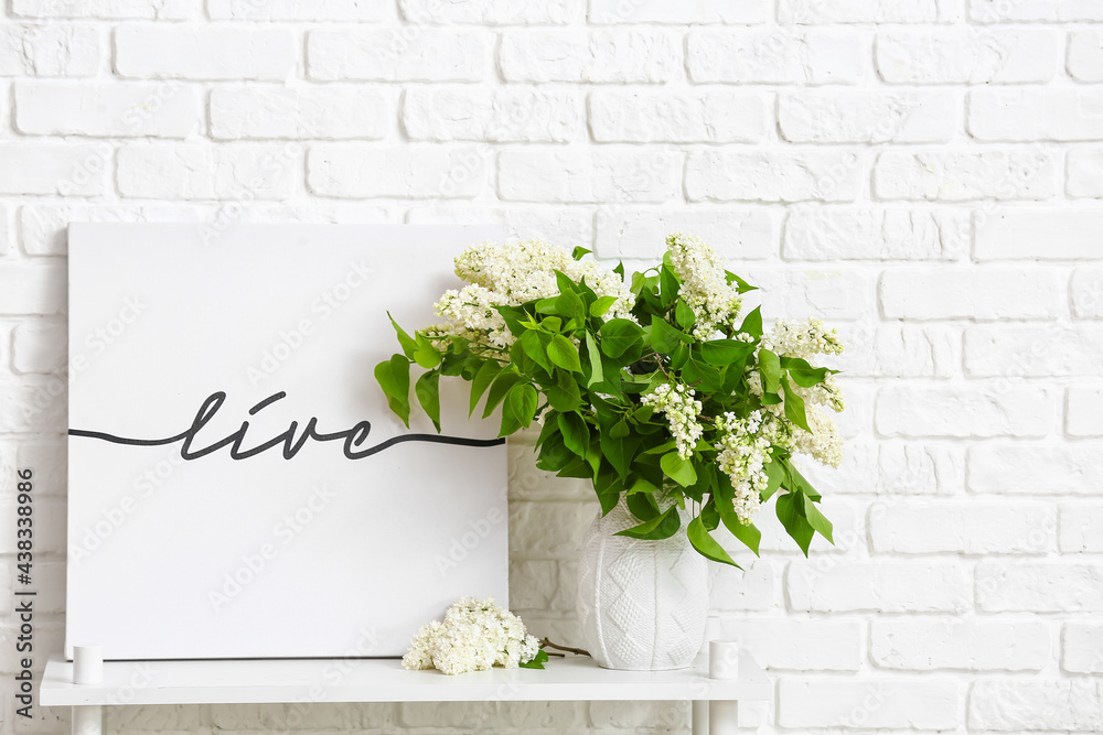 Vase with lilac flowers and picture on shelf near brick wall