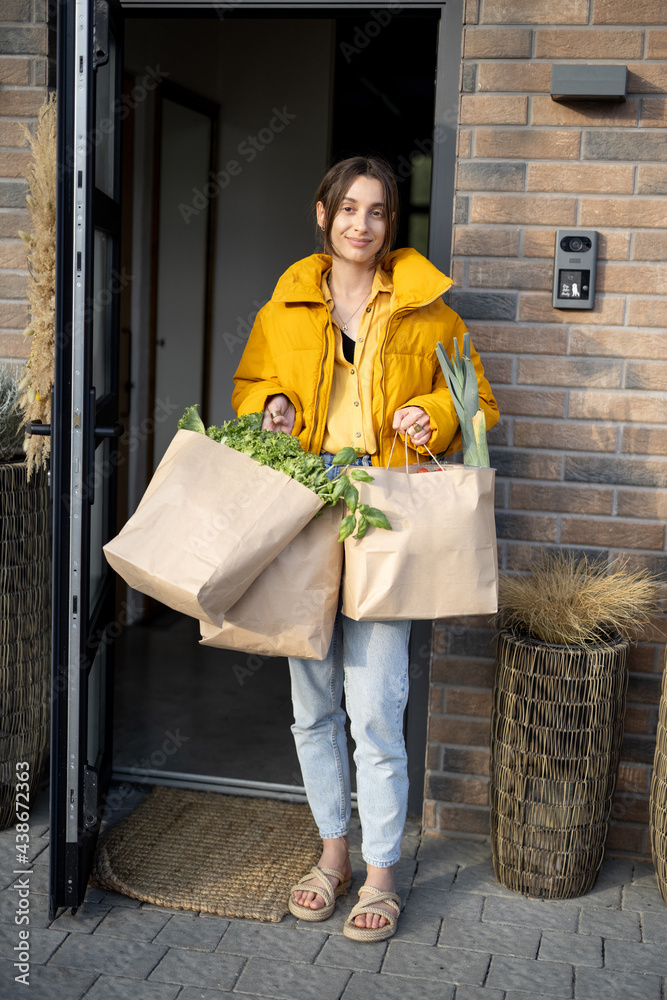 Young woman standing with paper bags full of fresh groceries on the porch of her house. Shopping hea