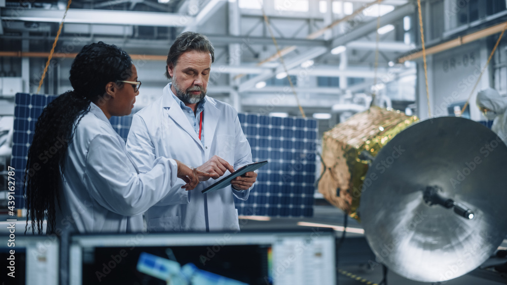 Two Engineers Talk, Use Tablet Computer while Working on Satellite Construction. Aerospace Agency Ma