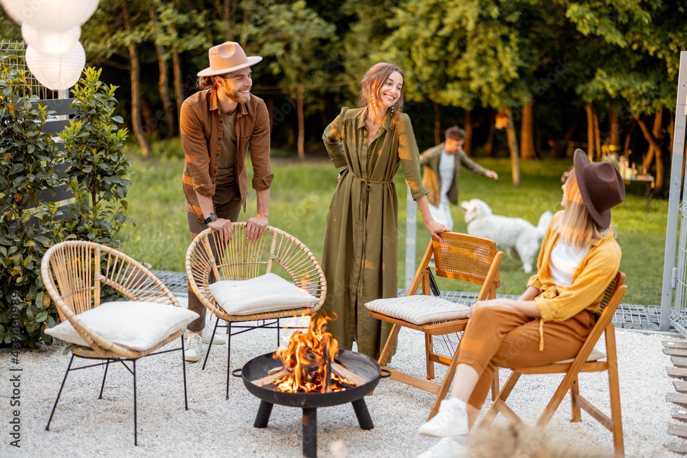 Friends sitting by a fireplace, paying with dog, having great summertime at backyard near the forest