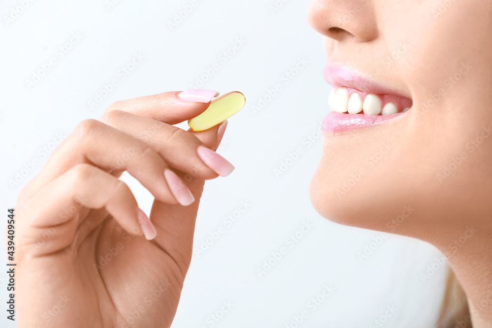 Young woman taking fish oil pill on light background, closeup