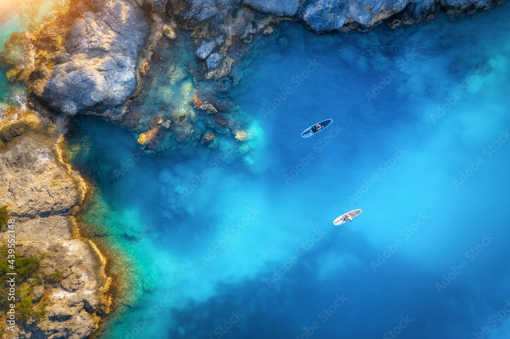 Aerial view of people on floating sup boards on blue sea, rocks, trees at sunset in summer. Blue lag