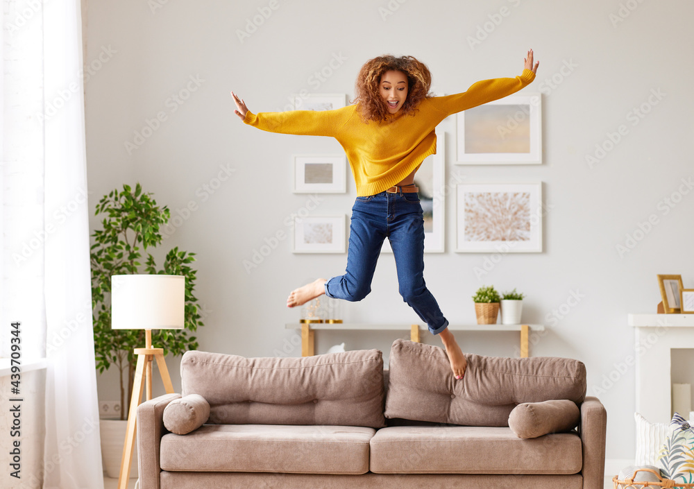 Happy african american teen girl jumping on sofa while having fun on weekend at home