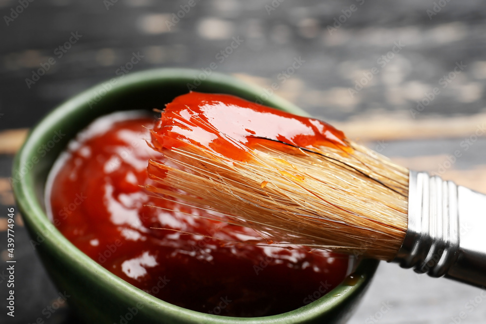 Bowl with tasty barbecue sauce and brush on dark wooden background, closeup