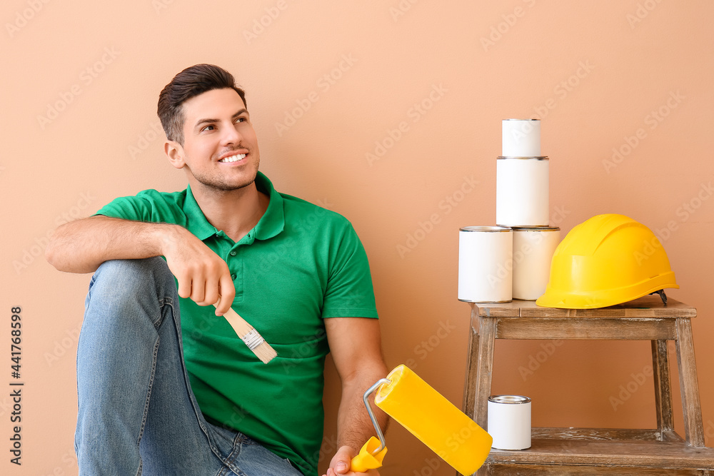 Young man with roller, brush and cans of paint on color background