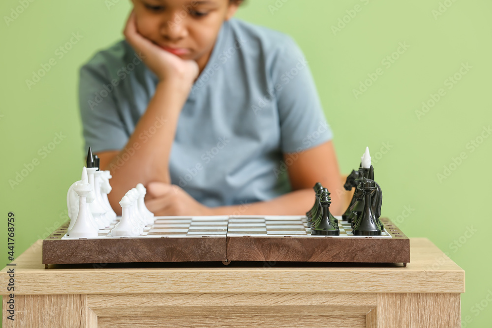 Cute African-American boy playing chess on color background