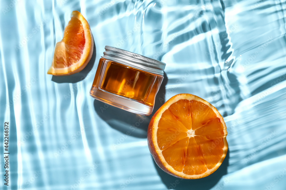 Jar of cosmetic product and cut orange in water on color background, closeup