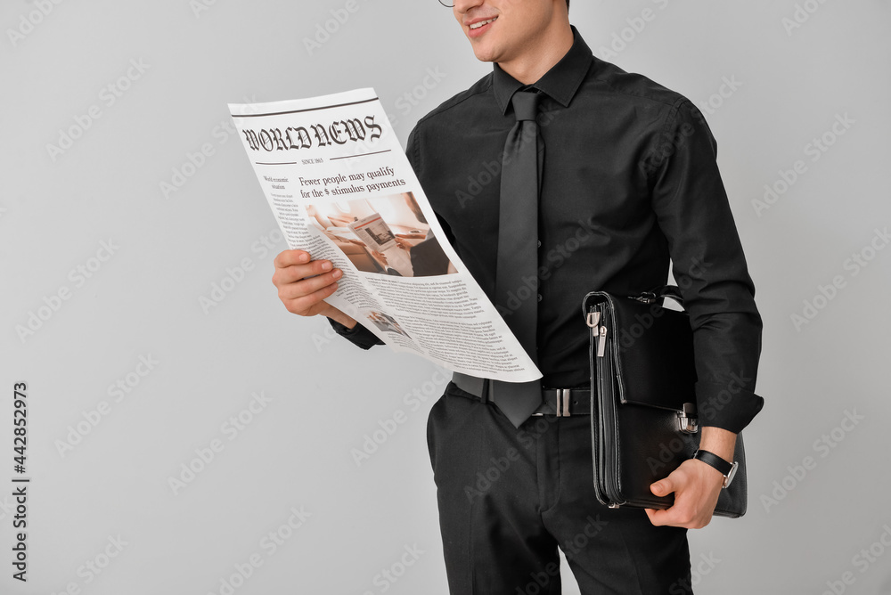 Young businessman with briefcase reading newspaper on light background